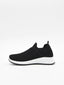 Zapatilla slip-on negra de punto knit con suela blanca y detalles en relieve en el tejido, sin cordones.